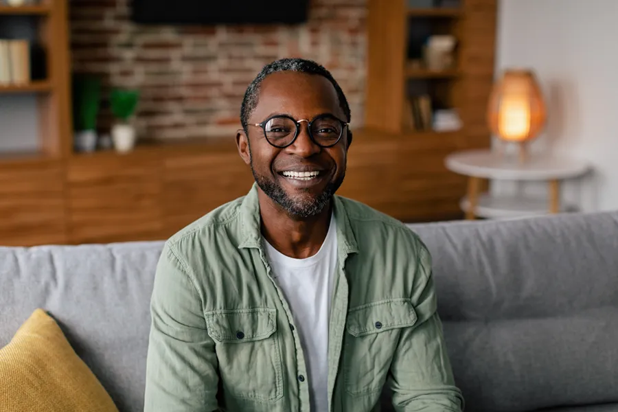 Andropause-Doctor A middle aged black man in a green button up shirt sits on his couch smiling, happy with his treatment for andropause and low testosterone from Markham Natural Health Centre in Greater Toronto.