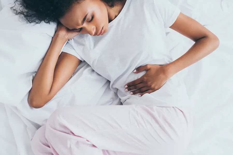 Crohns-Disease-Clinic A dark-skinned woman in white clothes lays on her bed clutching her middle before getting treatment for Crohn's Disease from Markham Natural Health Centre in Greater Toronto.