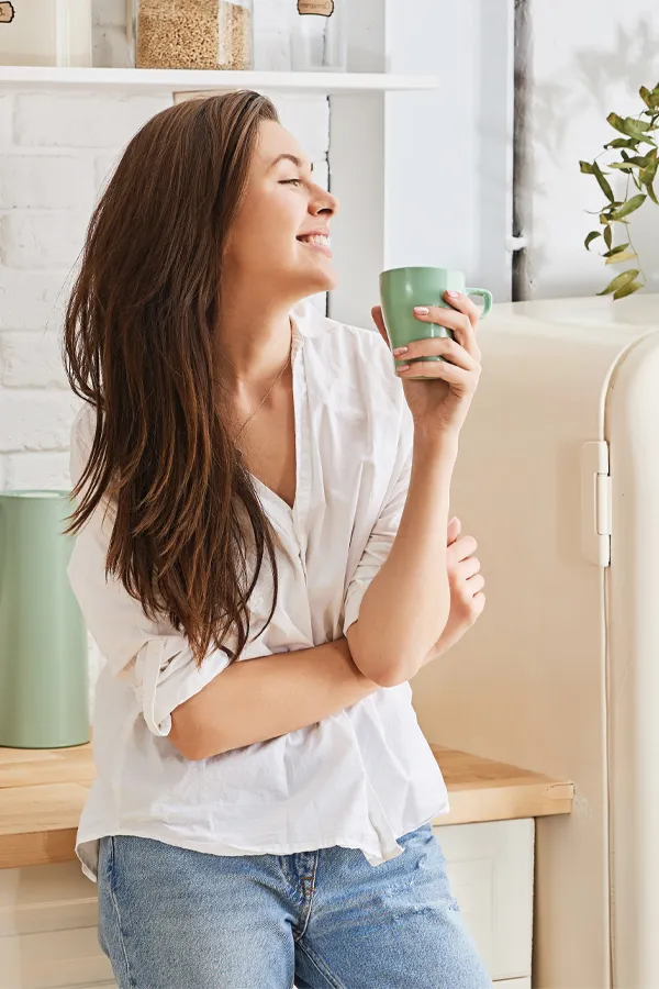 Crohns-Disease-Treatment A woman with a white blouse an jeans drinking tea in her kitchen, smiling after getting treatment for Crohn's Disease from Markham Natural Health Centre in Greater Toronto.