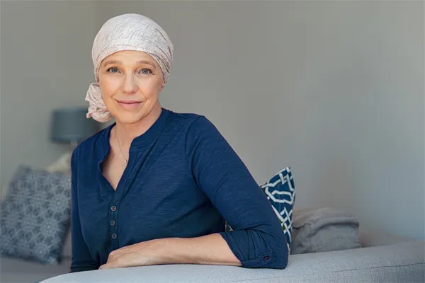 Functional-Oncology-Doctor A woman wearing a blue shirt with a white scarf tied around her head sitting on a couch and smiling at the camera, representing sensitive functional oncology treatment from Markham Natural Health Centre in Greater Toronto.