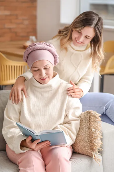 Functional-Oncology-Treatment A woman with a scarf tied around her head sits on a couch reading while another woman sits behind her with her hands on her shoulders warmly, representing sensitive functional oncology treatment from Markham Natural Health Centre in Greater Toronto.