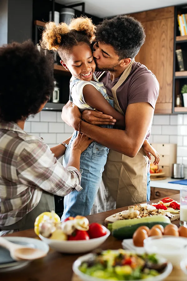 Gut-Health-Treatment A man holds his daughter and kisses her cheek as the family makes a healthy dinner in the kitchen. Get help for digestive issues and gut health from Markham Natural Health Centre in Greater Toronto.