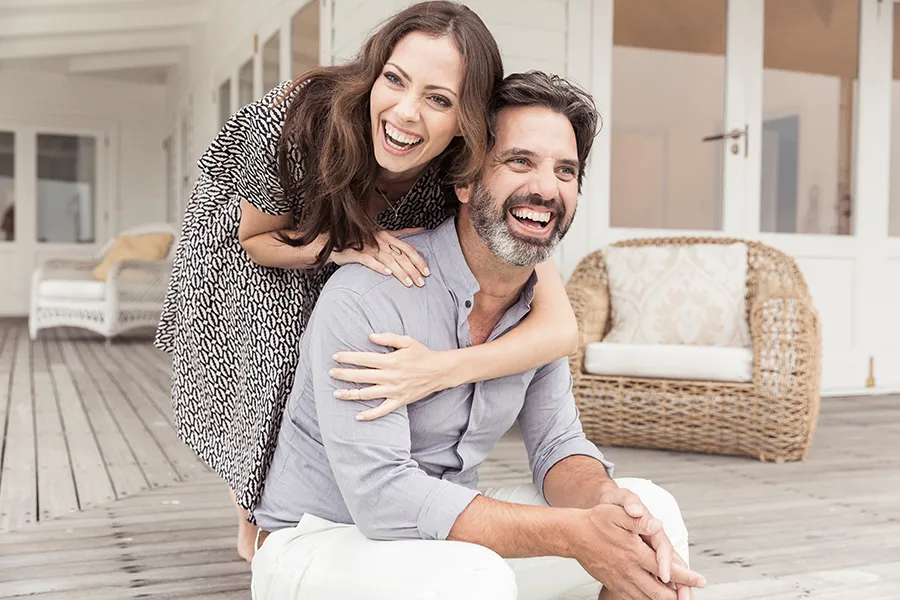 Health-and-Wellness-Services-Doctor A middle-aged couple on a wooden porch with wicker furniture; the woman is playfully hugging the man from behind. They are benefiting from the health and wellness services from Markham Natural Health Centre in Greater Toronto.