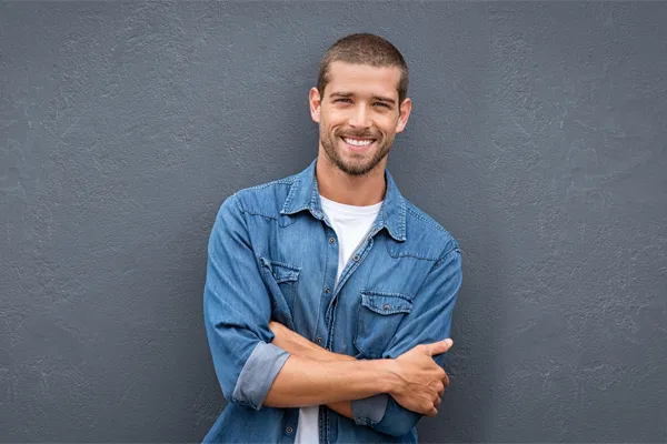 Hormones-for-Men-Doctor A man in a denim shirt stands smiling against a gray-blue wall, pleased with his testosterone hormone therapy from Markham Natural Health Centre in Greater Toronto.