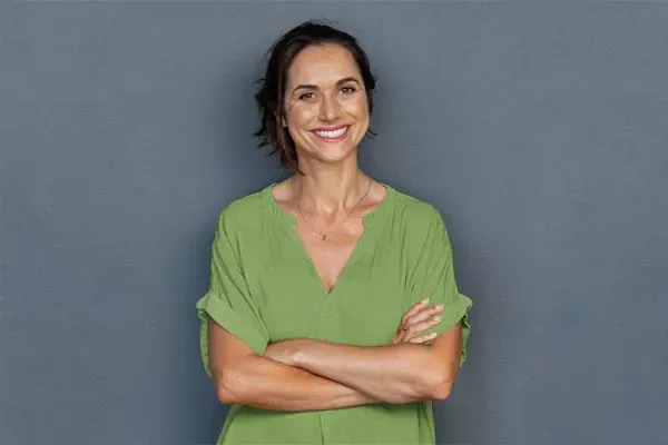 Hormones-for-Women-Doctor A woman in a green shirt stands smiling against a gray-blue wall, pleased with her hormone therapy from Markham Natural Health Centre in Greater Toronto.