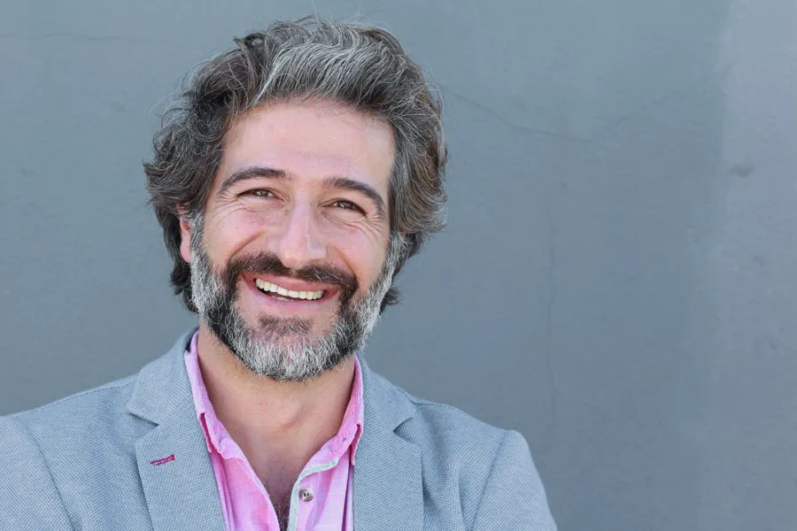 Hypogonadism-Clinic A smiling man with salt and pepper hair and beard wearing a pink shirt and gray blazer stands agains a gray wall. Get hypogonadism treatment from Markham Natural Health Centre in Greater Toronto.