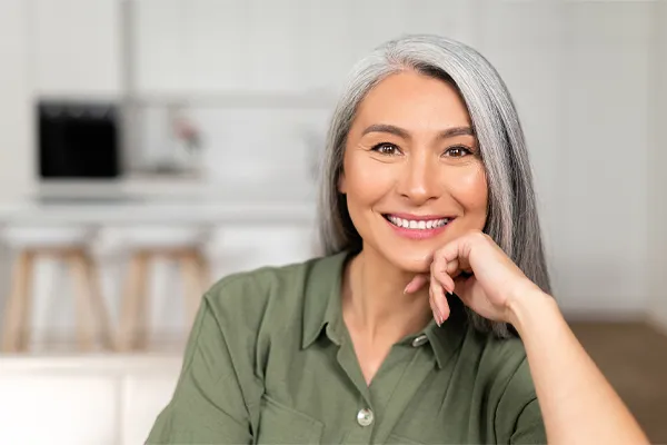Menopause-Clinic An asian woman with long gray hair and a green shirt sitting and smiling on her couch, happy with her menopause care from [NAME} in Greater Toronto.