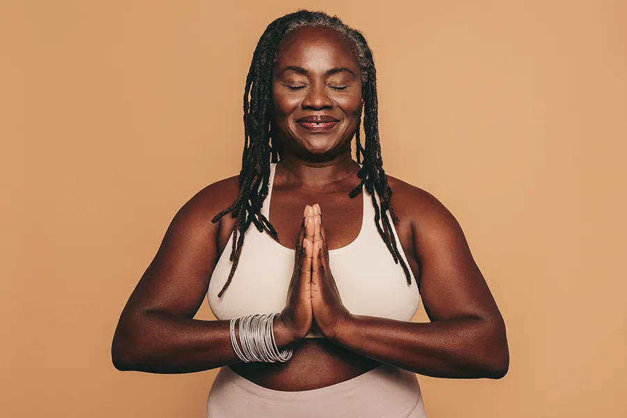 Obesity-Clinic An older African American woman confidently posing against a warm background smiling with hands in a prayer pose. Markham Natural Health Centre treats obesity using functional medicine in Greater Toronto.