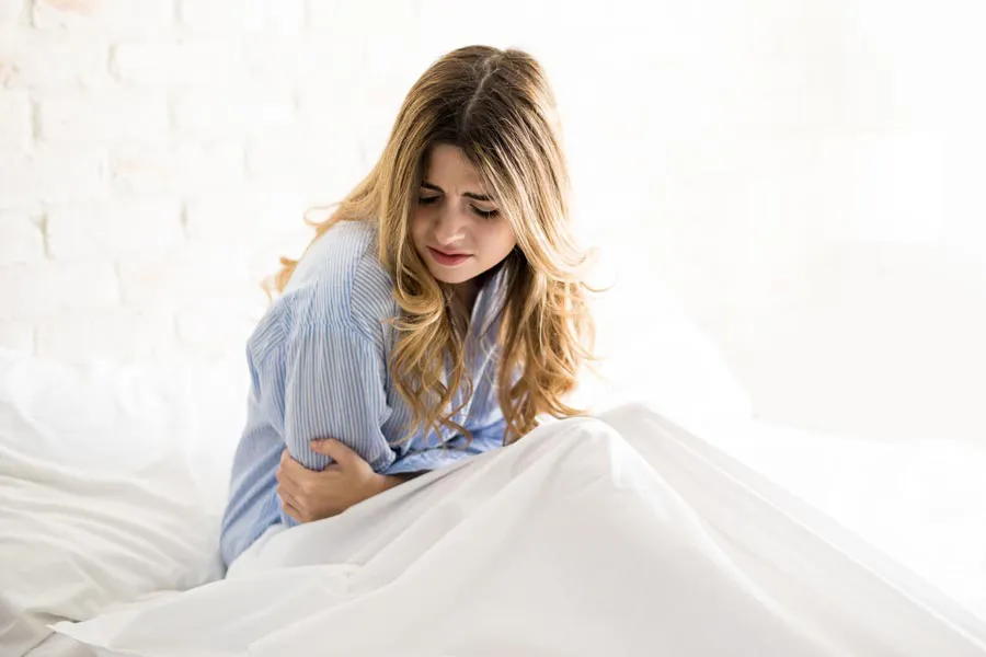 PMS-Therapy A woman with long blonde hair sits in her bed holding her stomach in pain, considering getting care for PMS from Markham Natural Health Centre in Greater Toronto.