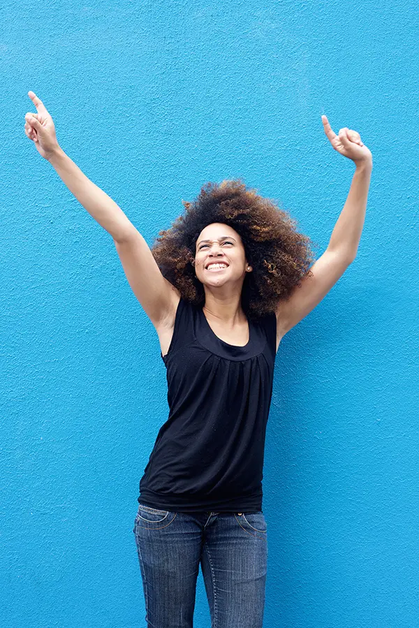 PMS-Treatment A woman in a dark blue tank top standing in front of a bright blue wall, raising her arms in celebration of relief from PMS from Markham Natural Health Centre in Greater Toronto.