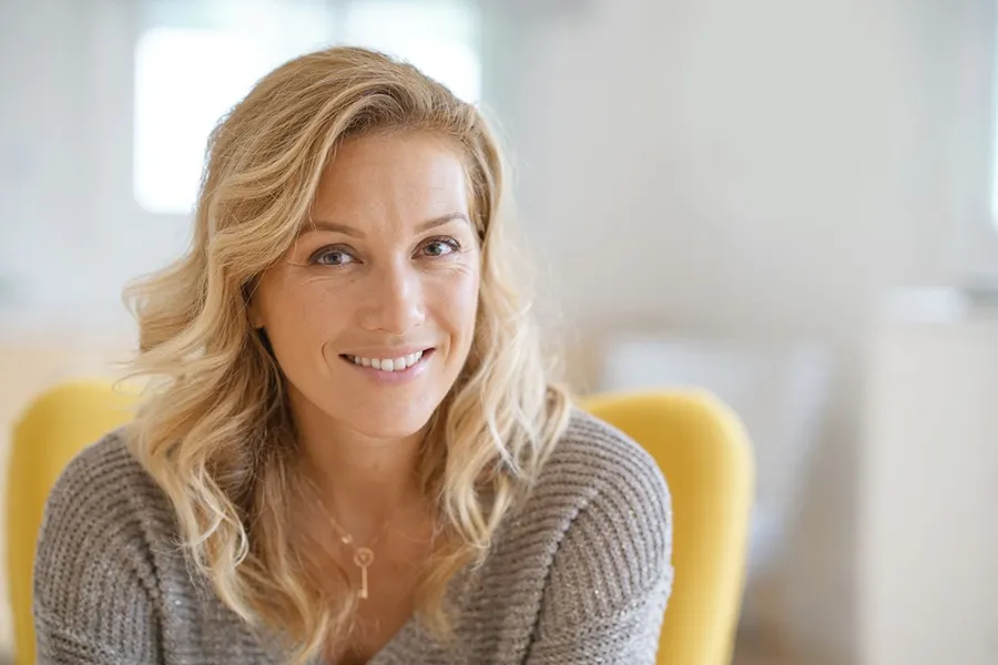 Perimenopause-Doctor A blonde woman wearing a a gray sweater sits inside in a yellow chair, smiling and happy with her perimenopause treatment from Markham Natural Health Centre in Greater Toronto.