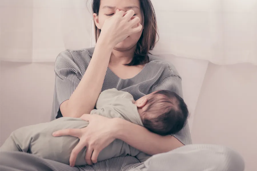 Postpartum-Hormone-Imbalance-Clinic A woman nursing an infant on couch rubs her eyes with exhaustion. Get treatment for postpartum hormone imbalance from Markham Natural Health Centre in Greater Toronto.